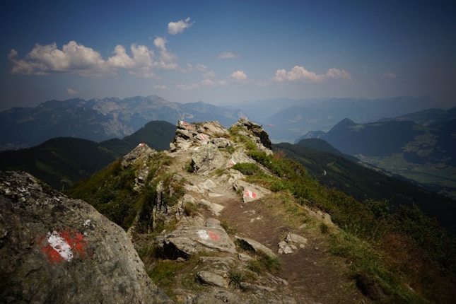 A scenic mountain trail winds across rocky terrain with patches of green grass and wildflowers. The trail is marked with red and white paint, suggesting a hiking route. In the distance, a vast expanse of rolling hills and valleys can be seen under a partly cloudy sky.