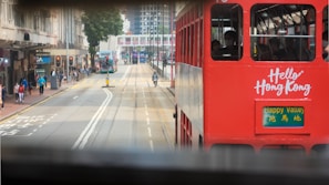 A red tram with the words 'Hello Hong Kong' on its side is traveling along a city street. The destination sign reads 'Happy Valley' in English and Chinese. Buildings line the street, and there are pedestrians walking and crossing the road. Traffic signals and road markings are visible.