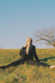 A person wearing a black suit sits on grass with legs stretched out on a sunny day. The individual is looking upwards, and a leafless tree is visible in the background against a clear blue sky.