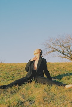 A person wearing a black suit sits on grass with legs stretched out on a sunny day. The individual is looking upwards, and a leafless tree is visible in the background against a clear blue sky.