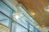 A modern interior scene featuring pendant lights hanging from a wooden ceiling. Large glass windows are present, allowing natural light to illuminate the space. The environment suggests an industrial or minimalist design, with visible air conditioning vents integrated into the wooden ceiling.