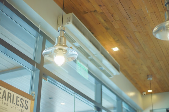 A modern interior scene featuring pendant lights hanging from a wooden ceiling. Large glass windows are present, allowing natural light to illuminate the space. The environment suggests an industrial or minimalist design, with visible air conditioning vents integrated into the wooden ceiling.