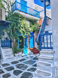 A charming, narrow alleyway with white stone steps and walls, accented by bright blue doors, windows, and railings. Lush green plants climb the walls, adding a touch of nature. The cobblestone path and vibrant floral arrangements enhance the quaint Mediterranean feel.