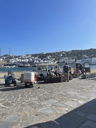 A small open-air market setup by a waterfront in a coastal town. Vendors are sitting beside vehicles and selling produce from crates. The backdrop includes blue water, moored boats, and white houses cascading down a hillside under a clear blue sky.