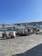 A small open-air market setup by a waterfront in a coastal town. Vendors are sitting beside vehicles and selling produce from crates. The backdrop includes blue water, moored boats, and white houses cascading down a hillside under a clear blue sky.