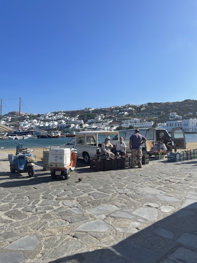 A small open-air market setup by a waterfront in a coastal town. Vendors are sitting beside vehicles and selling produce from crates. The backdrop includes blue water, moored boats, and white houses cascading down a hillside under a clear blue sky.