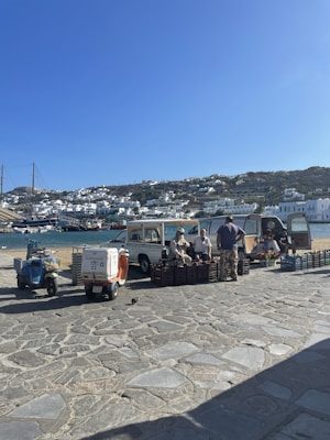 A small open-air market setup by a waterfront in a coastal town. Vendors are sitting beside vehicles and selling produce from crates. The backdrop includes blue water, moored boats, and white houses cascading down a hillside under a clear blue sky.