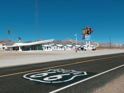 A retro-style motel with a large sign reading 'Roy's Vacancy Motel Cafe' is located along a desert road. Several palm trees and a modern, angular building are present in the foreground, with low, white buildings extending backward. Mountains can be seen in the background under a clear blue sky. The iconic Route 66 insignia is painted on the road in the foreground.