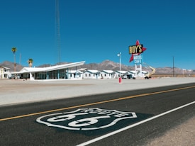 A retro-style motel with a large sign reading 'Roy's Vacancy Motel Cafe' is located along a desert road. Several palm trees and a modern, angular building are present in the foreground, with low, white buildings extending backward. Mountains can be seen in the background under a clear blue sky. The iconic Route 66 insignia is painted on the road in the foreground.