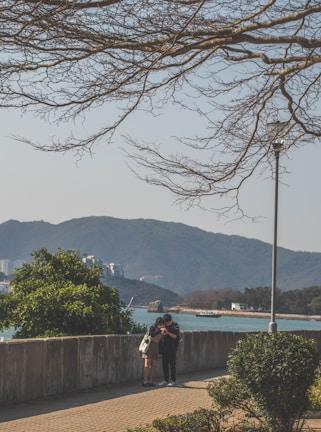 A couple stands closely together, looking at a smartphone in a serene outdoor setting near a waterfront. The background features distant mountains and a calm body of water. Leafless tree branches extend across the top portion of the image.