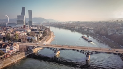 An urban landscape features a river flowing through a city, with a bridge spanning across it. Modern skyscrapers and industrial buildings are situated on one side, while residential areas with houses and streets are on the other. A boat travels along the river, and several cars move along the bridge. The image captures the blend of nature and urban life, with a backdrop of distant hills under a hazy sky.
