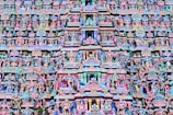 The image is a highly intricate and colorful facade of a Hindu temple, featuring numerous statues and carvings of deities and mythical figures. The architecture is elaborate, with vibrant shades of pink, blue, yellow, and green. The carvings are layered and densely packed, creating a rich tapestry of religious and cultural symbolism.