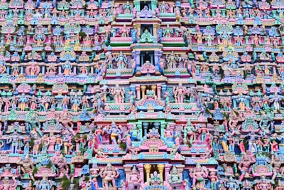 The image is a highly intricate and colorful facade of a Hindu temple, featuring numerous statues and carvings of deities and mythical figures. The architecture is elaborate, with vibrant shades of pink, blue, yellow, and green. The carvings are layered and densely packed, creating a rich tapestry of religious and cultural symbolism.
