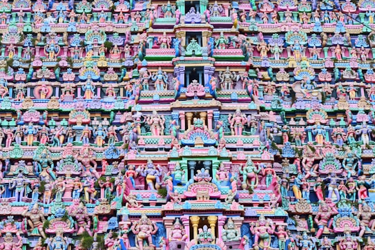The image is a highly intricate and colorful facade of a Hindu temple, featuring numerous statues and carvings of deities and mythical figures. The architecture is elaborate, with vibrant shades of pink, blue, yellow, and green. The carvings are layered and densely packed, creating a rich tapestry of religious and cultural symbolism.