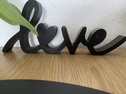 A wooden table surface is visible with a black decorative script that spells 'love'. A green leaf partially extends over the left side of the script. The background features a textured white wall.