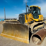 A large yellow bulldozer is parked on a dirt construction site under a blue sky. The machine's massive blade is prominently displayed, and a large cylindrical pipe lies on the ground beside it. In the background, several construction vehicles and equipment can be seen.