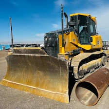 A large yellow bulldozer is parked on a dirt construction site under a blue sky. The machine's massive blade is prominently displayed, and a large cylindrical pipe lies on the ground beside it. In the background, several construction vehicles and equipment can be seen.