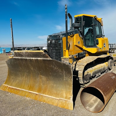 A large yellow bulldozer is parked on a dirt construction site under a blue sky. The machine's massive blade is prominently displayed, and a large cylindrical pipe lies on the ground beside it. In the background, several construction vehicles and equipment can be seen.