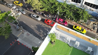 Aerial view of a street with multiple cars and motorbikes. The cars are in different colors including yellow, red, silver, and pink, parked alongside the road. Motorbikes are also visible on the street. The image shows a section of a building with a green rooftop garden and some chairs. Trees line the right side of the street, providing some greenery.