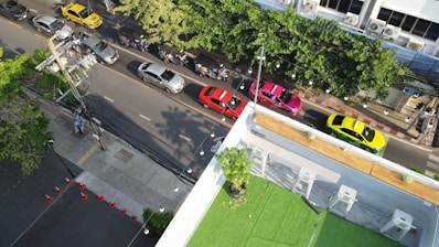 Aerial view of a street with multiple cars and motorbikes. The cars are in different colors including yellow, red, silver, and pink, parked alongside the road. Motorbikes are also visible on the street. The image shows a section of a building with a green rooftop garden and some chairs. Trees line the right side of the street, providing some greenery.