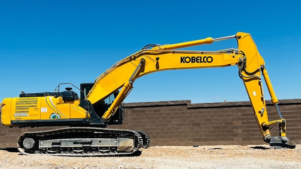 A large yellow excavator with the brand name visible on its arm is parked on a construction site. The machine has a long arm and is resting on a dirt ground in front of a brown brick wall under a clear blue sky.