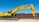 A large yellow excavator with the brand name visible on its arm is parked on a construction site. The machine has a long arm and is resting on a dirt ground in front of a brown brick wall under a clear blue sky.