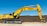 A large yellow excavator with the brand name visible on its arm is parked on a construction site. The machine has a long arm and is resting on a dirt ground in front of a brown brick wall under a clear blue sky.