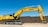 A large yellow excavator with the brand name visible on its arm is parked on a construction site. The machine has a long arm and is resting on a dirt ground in front of a brown brick wall under a clear blue sky.