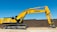 A large yellow excavator with the brand name visible on its arm is parked on a construction site. The machine has a long arm and is resting on a dirt ground in front of a brown brick wall under a clear blue sky.