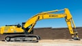 A large yellow excavator with the brand name visible on its arm is parked on a construction site. The machine has a long arm and is resting on a dirt ground in front of a brown brick wall under a clear blue sky.