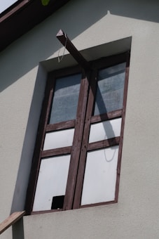 A window with wooden frames set into a light-colored concrete wall. The glass panes are opaque, suggesting frosted or textured glass, and a beam extends from above the window creating a shadow on the wall.