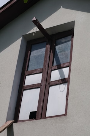 A window with wooden frames set into a light-colored concrete wall. The glass panes are opaque, suggesting frosted or textured glass, and a beam extends from above the window creating a shadow on the wall.