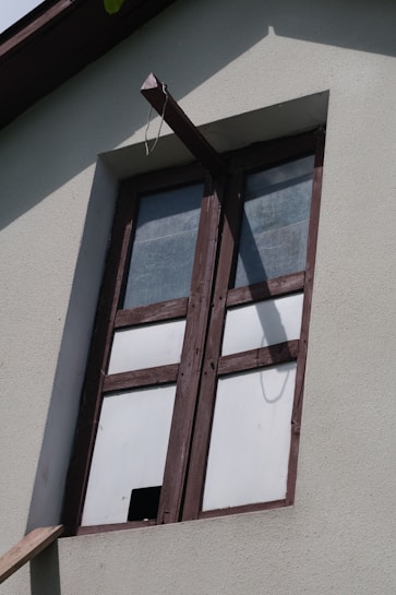 A window with wooden frames set into a light-colored concrete wall. The glass panes are opaque, suggesting frosted or textured glass, and a beam extends from above the window creating a shadow on the wall.
