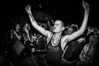 A monochrome photo captures a lively scene in a crowded venue. A person is at the forefront, displaying energetic body language with arms raised triumphantly, sporting a tank top and tattoos. Surrounding them, a group of people is enthusiastically engaged, some capturing the moment with their phones. The setting appears to be a concert or club.