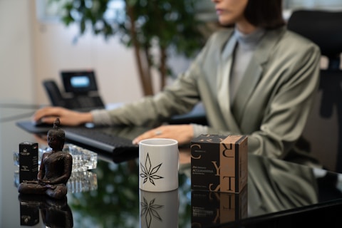 A person wearing a gray blazer is seated at a desk, typing on a keyboard. In the foreground, there is a white mug with a geometric design, a Buddha figurine, a crystal, and a brown box with black letters. A telephone can be seen in the background, and a plant adds greenery.