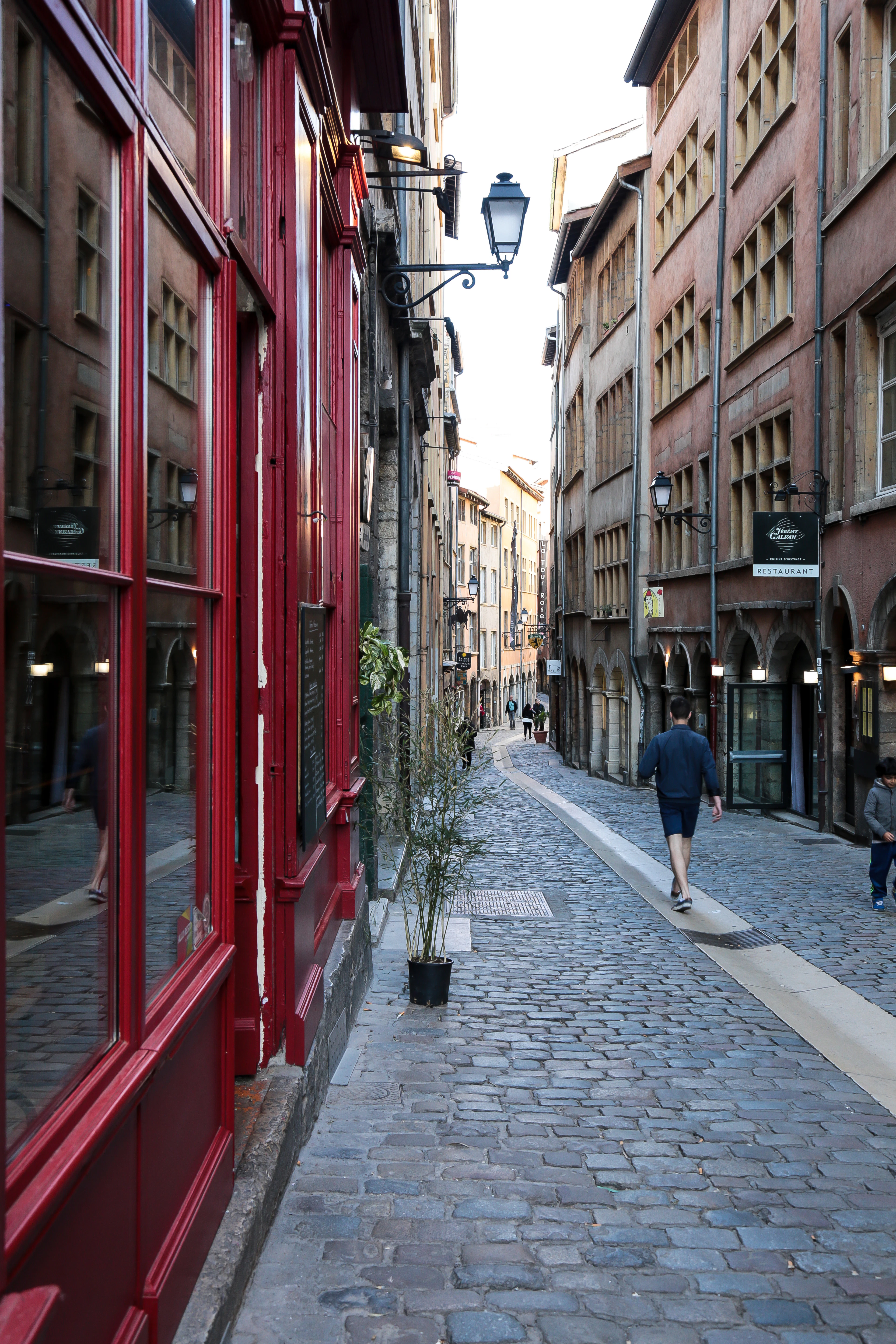 A cobbled lane in Vieux Lyon lined with Renaissance houses and arcaded ground floors, a deep red restaurant frontage on the left and a wrought-iron street lamp overhead.