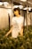 A woman stands in a room filled with cannabis plants, dressed in a white T-shirt with a graphic design and text 'Crystalweed High Quality CBD'. The environment appears to be an indoor cultivation area with artificial lighting and reflective surfaces.