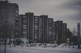 A row of tall, rectangular apartment buildings with numerous windows stands in an urban setting. In front of the buildings, there is a snowy landscape with a few bare trees and some signage with symbols and numbers. The sky appears overcast, giving the scene a gloomy appearance.