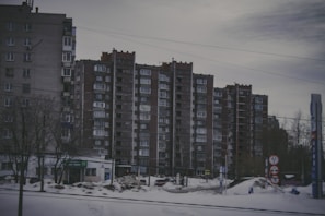 A row of tall, rectangular apartment buildings with numerous windows stands in an urban setting. In front of the buildings, there is a snowy landscape with a few bare trees and some signage with symbols and numbers. The sky appears overcast, giving the scene a gloomy appearance.