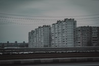 A group of large, rectangular, grey concrete apartment buildings seen in an urban setting. The sky is overcast, casting a gloomy atmosphere. In the foreground, there are railings and wires with string lights hanging across.