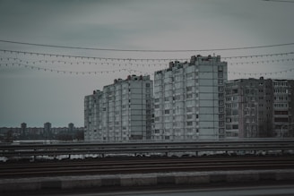 A group of large, rectangular, grey concrete apartment buildings seen in an urban setting. The sky is overcast, casting a gloomy atmosphere. In the foreground, there are railings and wires with string lights hanging across.