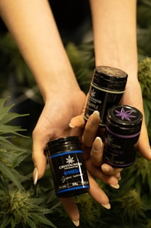A pair of hands holding three small black containers labeled with product names and surrounded by leafy green plants.