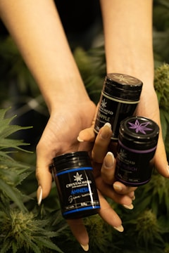 A pair of hands holding three small black containers labeled with product names and surrounded by leafy green plants.
