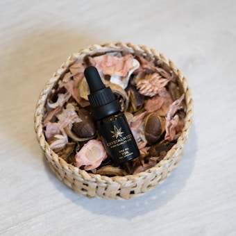 A bottle of CBD oil labeled 'Crystalweed' is set inside a wicker basket filled with dried flowers and petals. The basket is placed on a light-colored wooden surface.