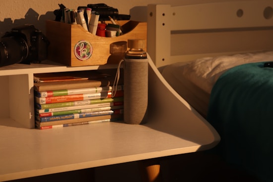 A tidy desk with a collection of multicolored books neatly stacked on the left, alongside a cylindrical speaker. On top of the desk, a wooden organizer holds a variety of pens, markers, and a camera. Warm sunlight casts shadows across the scene, creating a cozy, organized atmosphere.