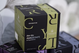 A box labeled 'Elixir Slim Amazzonite 10 pyramidal filters' with a design featuring large overlapping letters. The box is primarily green and black with the brand name 'Crystal Tea' and mentions 'high quality CBD'.
