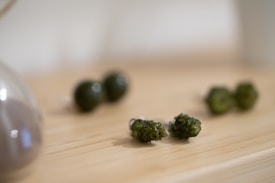 The image depicts a close-up of several small, textured, green stones placed on a light wooden surface. They have an uneven surface with a somewhat glossy finish. In the foreground, a few of these stones are in focus, while others are softly blurred in the background. An object with a reflective surface and soft curvature is partially visible to the left.