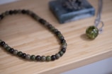 A beaded necklace made of greenish and dark-colored round stones is placed on a light wooden surface. Near the necklace, there is a spherical pendant attached to a silver chain, with the pendant displaying a green marbled texture. In the background is a square, bluish-green stone block.