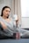 A person in comfortable loungewear is sitting on a couch holding a white mug with a black geometric design. A small box with text and logos is placed on the couch in front of them. The room has neutral-colored curtains and soft lighting.