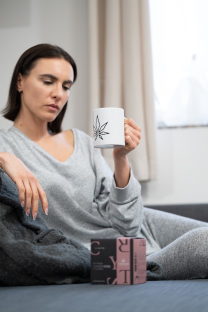 A person in comfortable loungewear is sitting on a couch holding a white mug with a black geometric design. A small box with text and logos is placed on the couch in front of them. The room has neutral-colored curtains and soft lighting.
